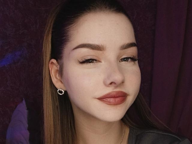 BitterPeach has light skin, brown eyebrows, long brown hair, and is wearing subtle eyeliner, a small earring, and red lipstick, smiling slightly.
