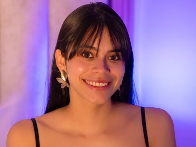 AlanaBlake has black hair, bangs, and a smile showing white teeth. She's wearing a black top with silver butterfly earrings, and her skin is a warm tone.