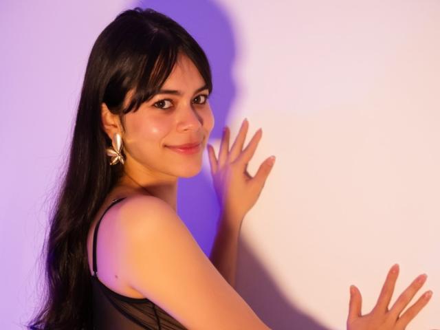 AlanaBlake with long black hair, smiling, wearing a black top, and gold earrings, has her hands and fingers against a white wall with purple lighting.