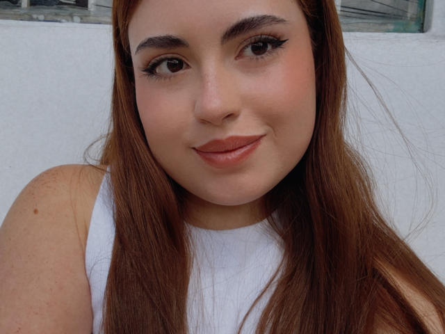 VicluX hat ein süßes Gesicht, braune Augen und langes, braunes Haar, trägt ein weißes Tanktop, hat volle Lippen und dezentes Make-up und lächelt sanft in die Kamera..