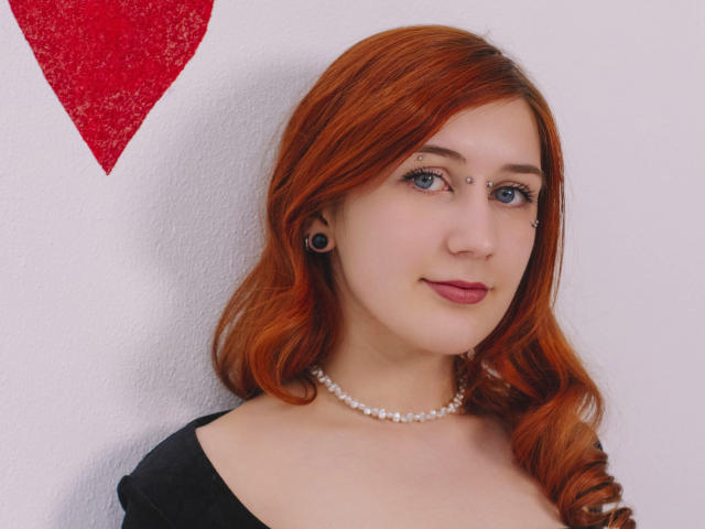 PollyFoxy has red hair, blue eyes, black ear gauges, nose ring, pearl necklace, and black top, with a subtle smile. Her tits are small, and she's cute.