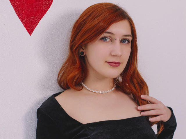 PollyFoxy with red hair, black off-shoulder top, pearl necklace, black ear gauges, subtle makeup, and a gentle smile, standing against a white wall with a red triangle.