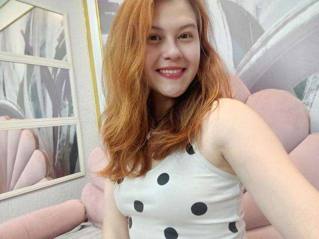 TaylorCarpert with red hair, smiling, wearing a white polka dot tank top, showing her tits, pink upholstered chair, mirror, and pink wall in the background.
