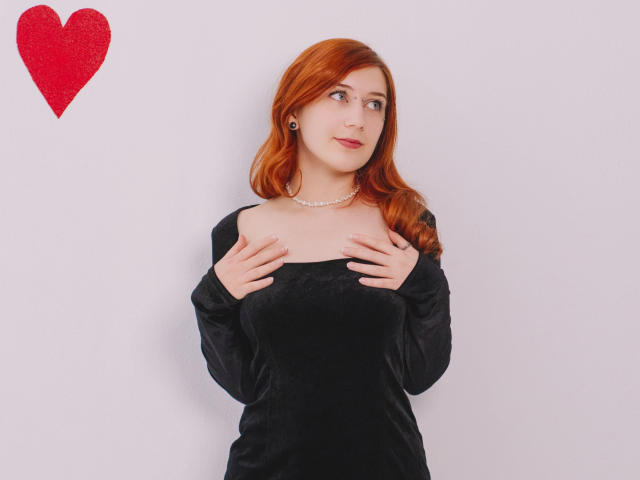PollyFoxy with red hair, wearing a black velvet dress, hands on chest, pearl necklace, looking up, red heart on wall, white background, cute, attractive.