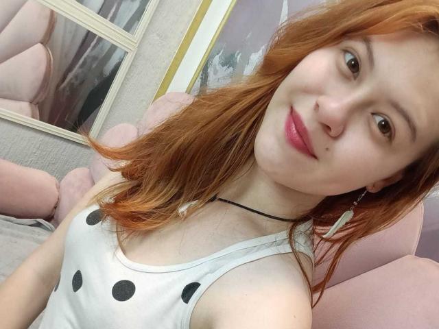 TaylorCarpert, a redhead with pink lips, wearing a white tank top with black polka dots, has small tits and a black choker, smiling in a selfie.