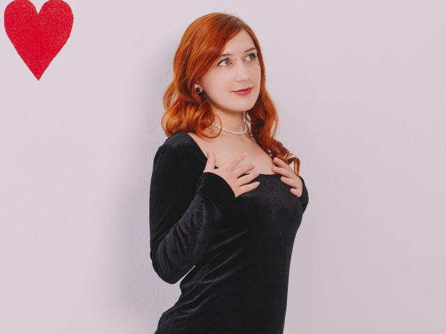 PollyFoxy with red hair, wearing a black velvet dress, hands on her tits, smiling, pearl necklace, red heart in top left, white background.