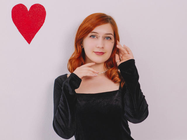 PollyFoxy with red hair, wearing a black velvet dress, touching her chin, pearl necklace, red glitter heart in top left, light skin, blue eyes, slight smile, looking at camera.