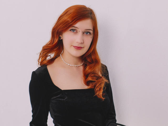 PollyFoxy has red hair, wearing a black velvet dress, pearl necklace, and small earrings, with a subtle smile, standing against a white wall.