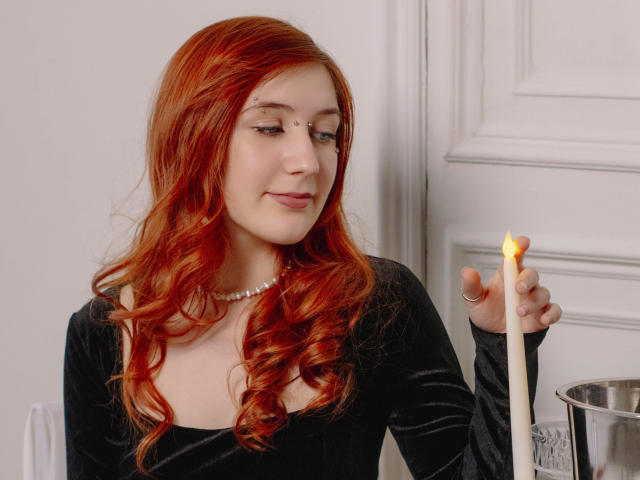 PollyFoxy with red hair, black velvet dress, pearl necklace, holding a burning white candle, smiling softly, in a silver cup.