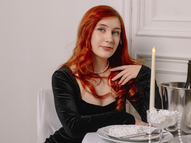 PollyFoxy with red hair, black velvet dress, pearl necklace, sitting at a table with a candle, white plate, silver bowl, and glass with pearls. Her tits are visible.