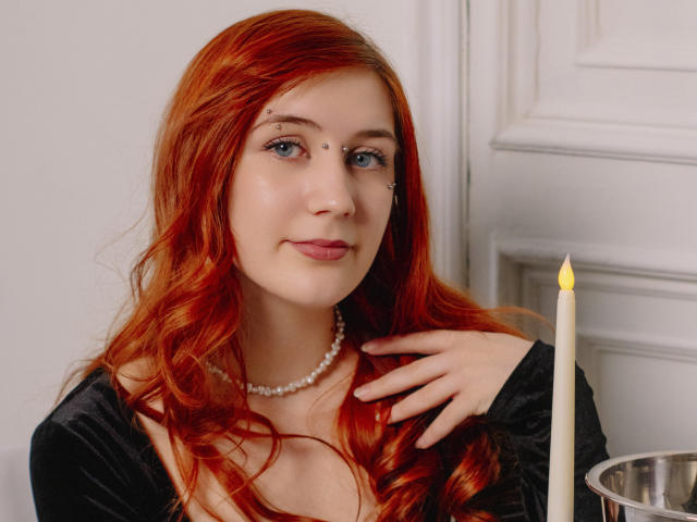PollyFoxy has red hair, blue eyes, and a pearl necklace, wearing a black top, with a lit candle in front of her. She's touching her chest with one hand.