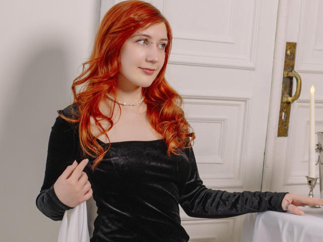 PollyFoxy with red hair, wearing a black velvet dress, pearl necklace, and black sleeves, stands near a white door, right hand on a table with a candle.