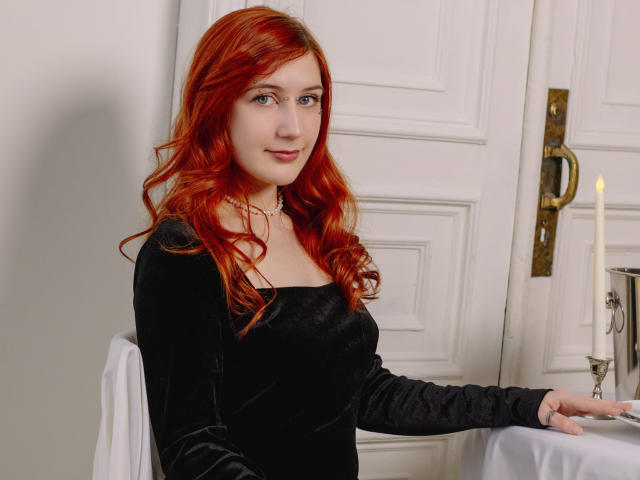 PollyFoxy with red hair, wearing a black velvet dress, sits by a white table with a candle, smiling softly, her hand resting on the table.