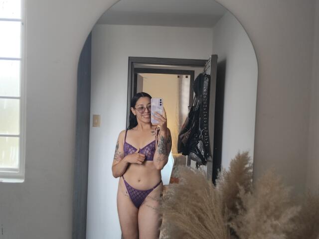 Mllevv stands in a mirror, taking a selfie with a pink phone. She wears a purple lace bra and matching thong, showing tattoos on her left arm and right thigh. She has dark hair, glasses, and a smile. The reflection shows a doorway with hanging clothes and potted pampas grass in the foreground. The image is a realistic photograph with natural lighting.