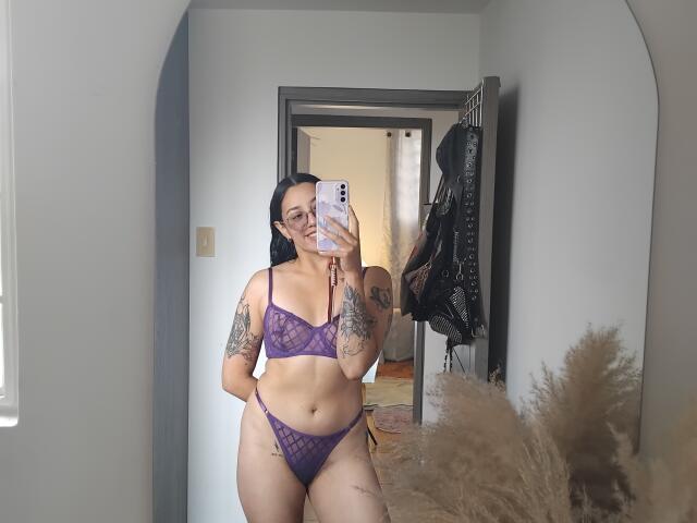 A mirror selfie shows Mllevv, a tattooed woman with long black hair, wearing a purple lace bra and matching thong. She holds a white phone in her right hand, partially covering her face. Her left arm, featuring a large tattoo, rests by her side. She has a curvy physique with visible tattoos on her right arm and thigh. Puffy beige pampas grass is in the lower right corner. The room has a gray door and black leather jacket on a hook in the background. Natural light from a window on the left.