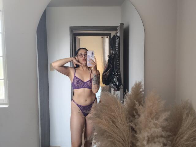 Mllevv stands in front of a mirror, taking a selfie with a phone. She's wearing a purple lace bra and matching thong. She has a tattoo on her left arm and is touching her hair with her right hand. She's light-skinned with long dark hair. The mirror has a dark frame, and there's a puffy brown grass-like plant in the bottom right. Mllevv's face is slightly turned, and she's looking at the phone. The image is casual and focused on her reflection.
