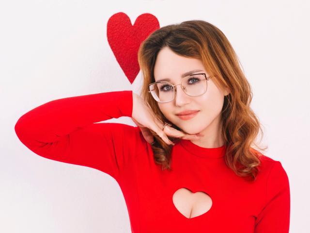 TeresaBirt has wavy, light brown hair and wears clear-framed glasses. She's in a red, long-sleeve shirt with a heart-shaped cutout revealing a small cleavage. She's touching her chin with her right hand and looking at the camera with a slight smile. A red heart is mounted on the white wall behind her. The composition is clean and focused on TeresaBirt, with a simple, white background that contrasts with her red shirt. The photograph has a soft, natural light that enhances her fair skin and subtle makeup. The style is modern and slightly casual.
