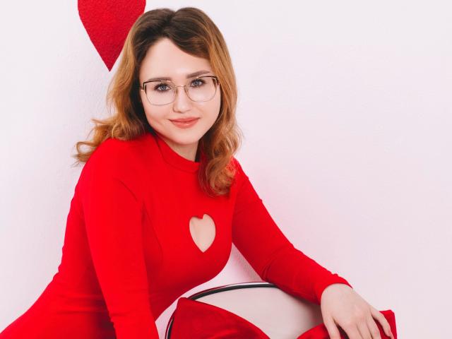 TeresaBirt with wavy brown hair, glasses, and red heart-cutout dress, sitting on red bag, smiling, red heart wall decoration, white background, sexy, cute, attractive.