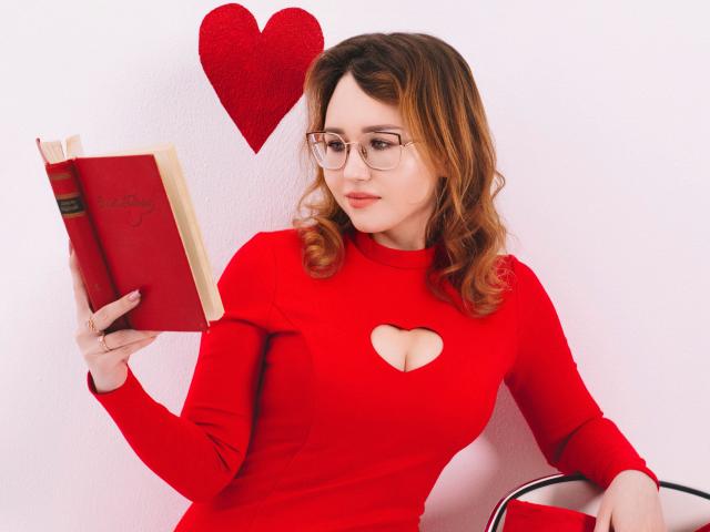 TeresaBirt with glasses, red dress, heart cutout, reading book, red heart wall decoration, wavy brown hair, sitting, right arm on chair, left hand holding book.