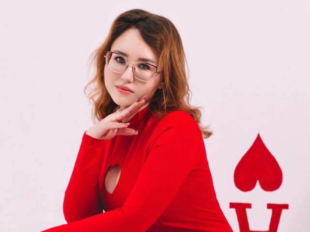 TeresaBirt with wavy brown hair, glasses, and a red long-sleeve top with a cutout, resting her chin on her hand, showing her ass with a playful smile.