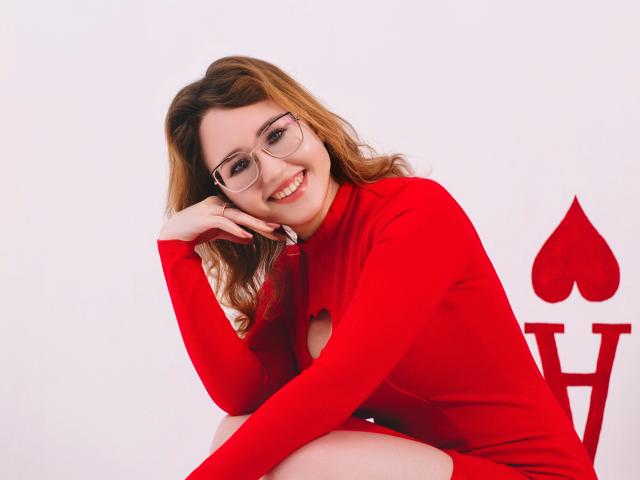 TeresaBirt, with glasses and red dress, smiling, sitting, hand on cheek, showing ass, brown wavy hair, white background, heart-shaped red chair.