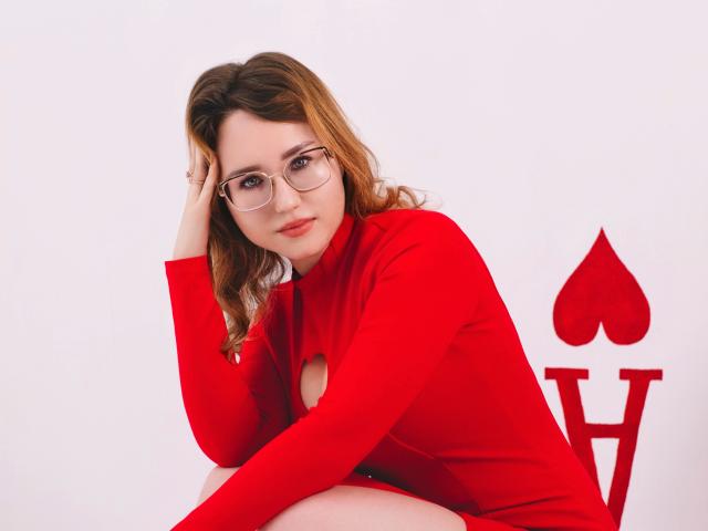 TeresaBirt with glasses, red dress, and cutout, sitting, hand on head, looking at camera, wavy brown hair, white background, red heart and spade symbols in the background.
