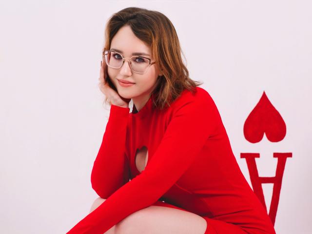 TeresaBirt with glasses, red dress, and cut-out, sitting with chin on hand, brown wavy hair, white background, wearing a cute and confident expression. Boobs visible through dress.