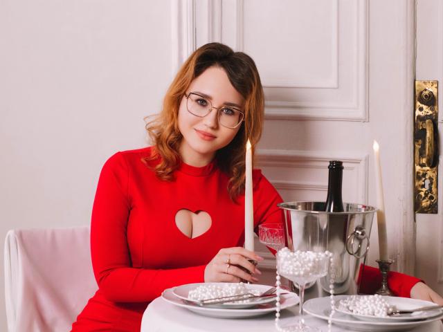 TeresaBirt with glasses, red dress, heart cutout, showing tits, holding candle, sitting at table with champagne, ice bucket, glasses, and pearl decorations, smiling softly.