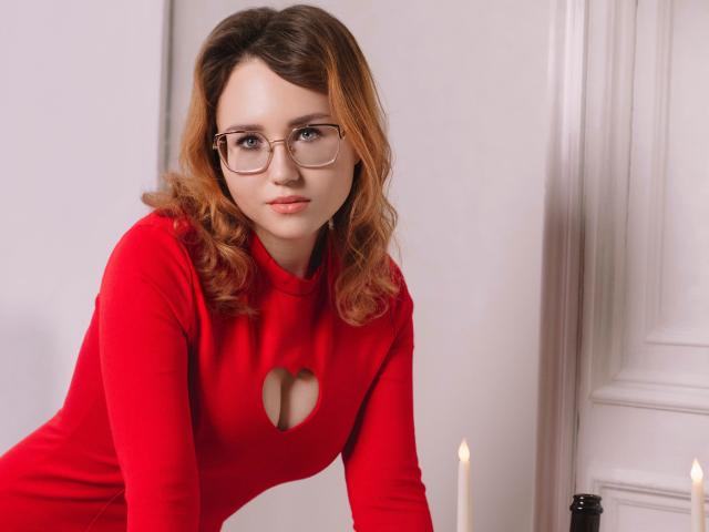 TeresaBirt with red cutout dress, glasses, and wavy brown hair, leaning forward, showing her small tits, looking serious. White candles in the background.