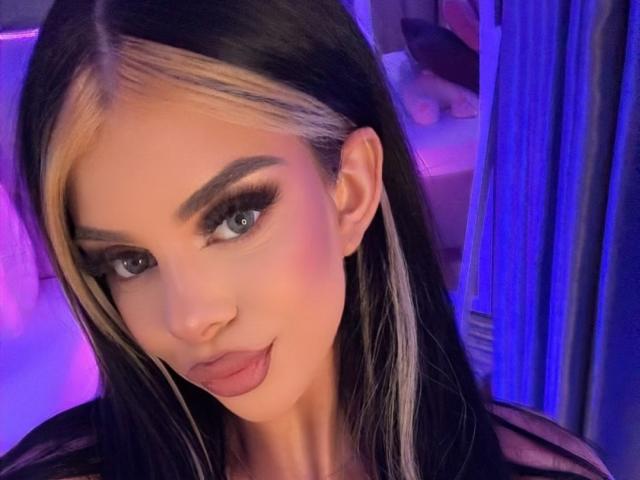 IvyRaven has dark black hair with blonde highlights, blue eyes, and is giving a slight pout with her lips. She's wearing heavy eyeliner and has a cute face.