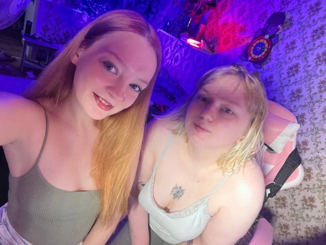 Two women with RadiantStwo: one with long red hair, small tits, green tank top, smiling; the other with short blonde hair, small tits, white tank top, neutral expression.