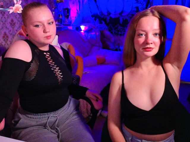 Two RadiantStwo girls, one with a nose ring, black mesh top, and gray pants, the other in a black crop top, posing in a blue-lit room.