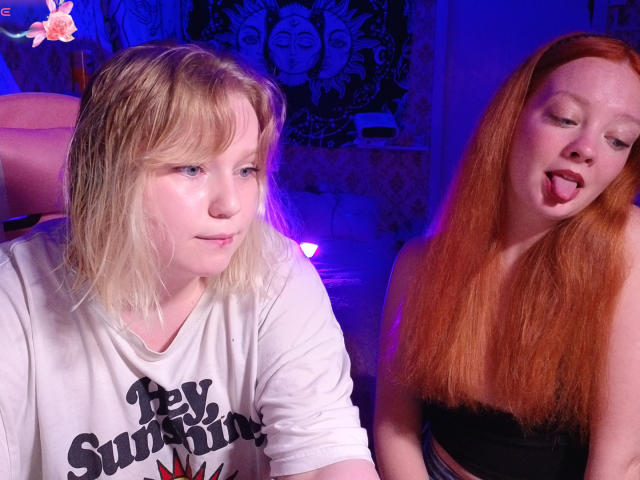 Two girls, RadiantStwo with blonde hair and small tits, wearing a white shirt, and another with red hair and a black top, tongue out, in a blue-lit room.