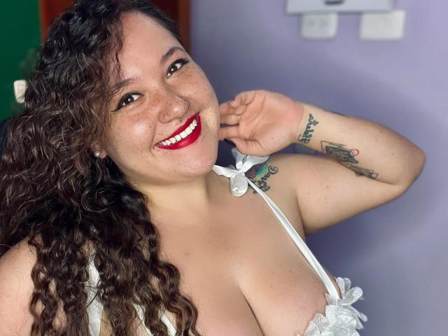 EmiliaMora with curly brown hair, red lipstick, smiling, white lace bra, visible large tits, tattoos on right arm, hand near face, light skin, purple wall background.