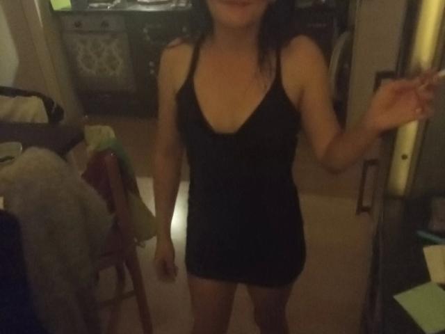 Elliefucku30-hot in a black, low-cut, sleeveless dress, showing her small tits and ass, with dark hair, standing in a dimly lit room, holding a door frame.