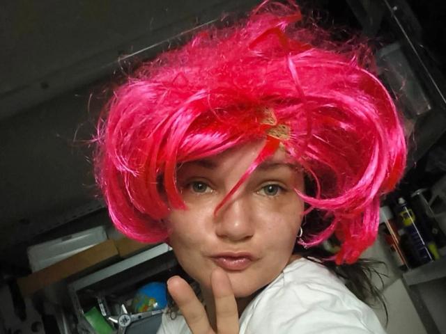Elliefucku30-hot with bright pink, curly wig, medium skin, green eyes, and white shirt, making a peace sign, showing slightly pouted lips. Shiny hair with red accent.