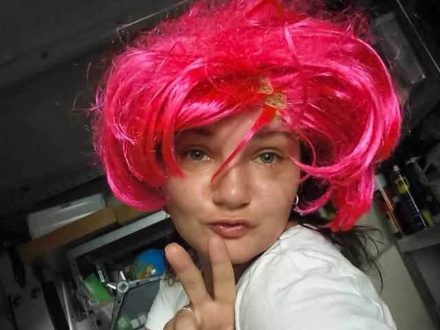 Elliefucku30-hot with bright pink, fluffy wig, green eyes, white shirt, and pink lipstick, making a peace sign, in a dimly lit room, showing a cute, playful expression.