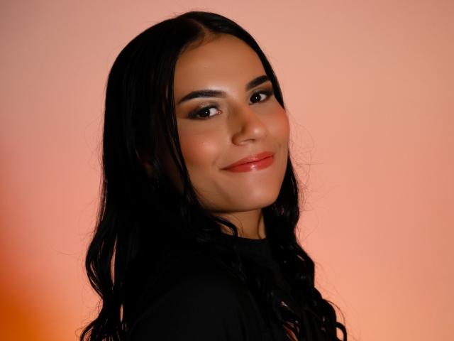 BellaTorres has dark, wavy hair, brown eyes, and a slight smile with red lips. She's wearing black and has a soft, warm glow on her face.
