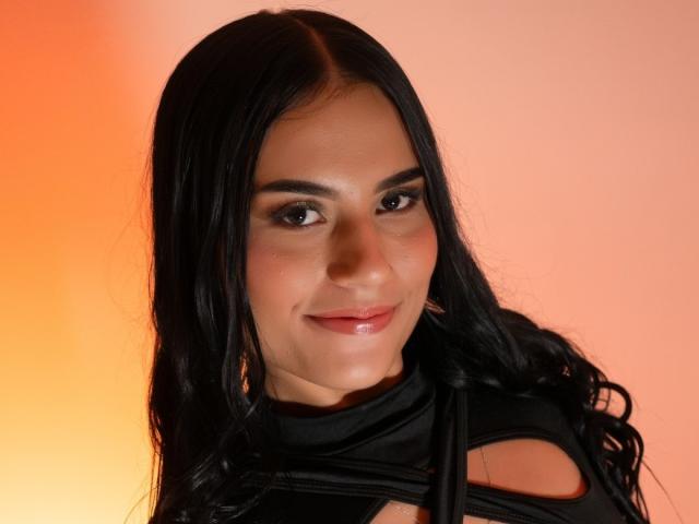 BellaTorres has long black hair, dark eyes, and a slight smile. She's wearing a black top with cutouts, showing a bit of her chest. Her skin is fair.