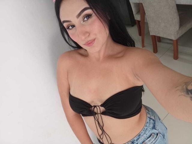 HiilaryDolce with black strapless top showing small tits, tied front, and blue jeans, smiling, long black hair, tattoo on right arm, standing against white wall.