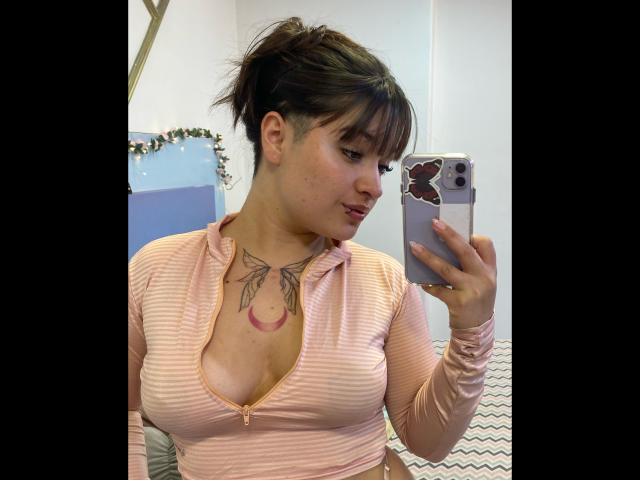 AnikaFleur with brown hair in a messy bun, pink striped top, zipper down, showing her tits and butterfly tattoo on her chest, taking a selfie with a butterfly phone case.