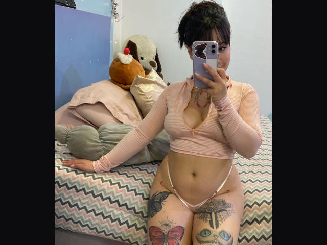 AnikaFleur with short black hair, pink top, and pink thong, showing her tits, tattoos on thighs, taking a mirror selfie, holding a phone, in a bedroom.
