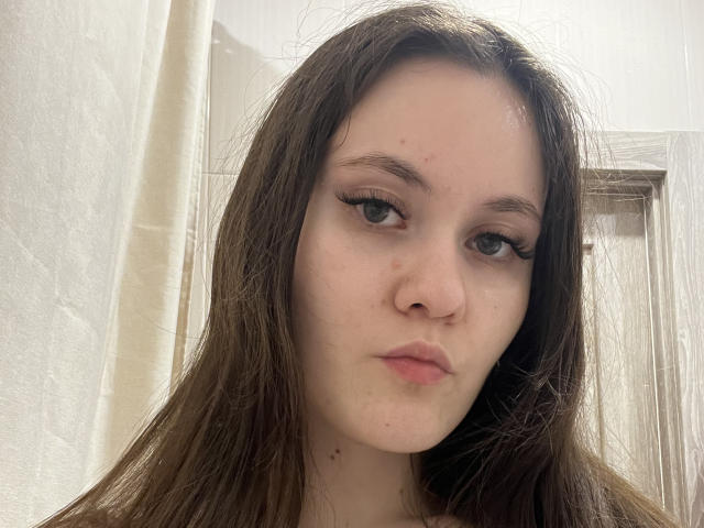 ClaudeGullett has long brown hair, light skin, and small black eyeliner. She has a pimple on her forehead, and her nose is slightly upturned. Her expression is neutral.