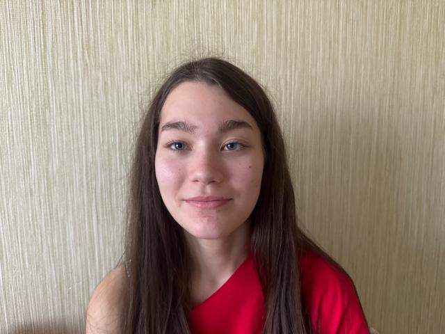 MerrieWirebaugh has long brown hair, blue eyes, and a slight smile, wearing a red shirt, with light skin and a few small acne spots on her face.