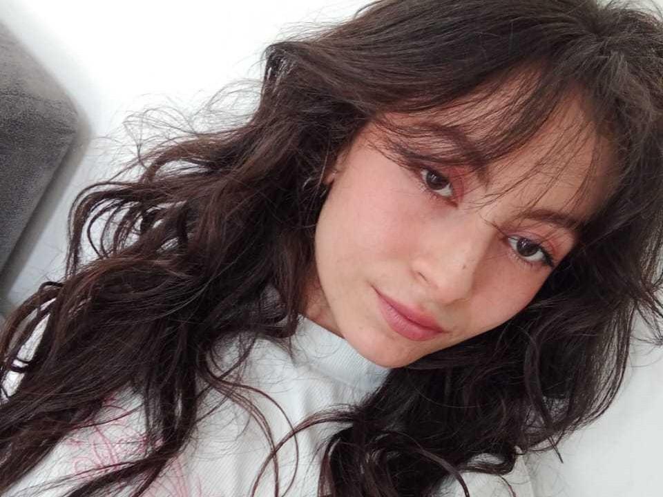 ZoeVelvet has long, wavy dark brown hair, pinkish skin, and brown eyes. She's wearing a white shirt with pink details, looking up at the camera with a slight smile.