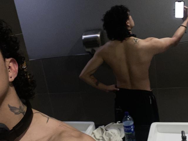 JayTaylor, ohne Hemd, mit lockigen schwarzen Haaren und Tätowierungen an Schulter und Hals, steht mit dem Rücken zur Spiegel, die Hände auf der schwarzen Hose, wobei die Spiegelung zu sehen ist. Eine Wasserflasche steht auf dem Waschbecken..