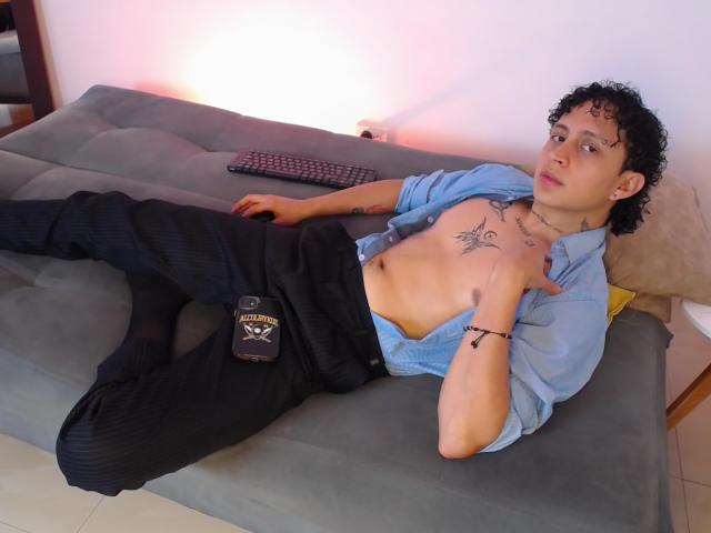 Jay Taylor, con i capelli neri ricci, camicia aperta che mostra tatuaggi sul petto, pantaloni neri, sdraiato su un letto grigio, che guarda la telecamera, mani sul petto, rilassato, casual, tatuato, leggermente muscoloso..