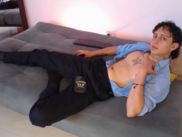 JayTaylor, torse nu, pantalon noir, chemise bleue, tatouages, allongé sur un canapé gris, jambes écartées, bras sur la poitrine, regardant la caméra, boxers noirs avec le logo "X", cheveux bouclés, peau claire..