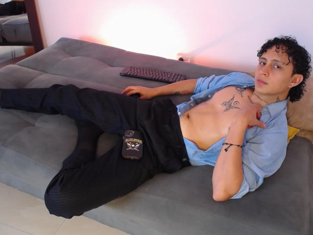 JayTaylor, torse nu, avec des cheveux noirs bouclés, allongé sur un canapé gris, portant un pantalon noir et une chemise bleue, avec des tatouages sur la poitrine, un bracelet noir, un clavier sur le canapé, une bouteille sur le pantalon..