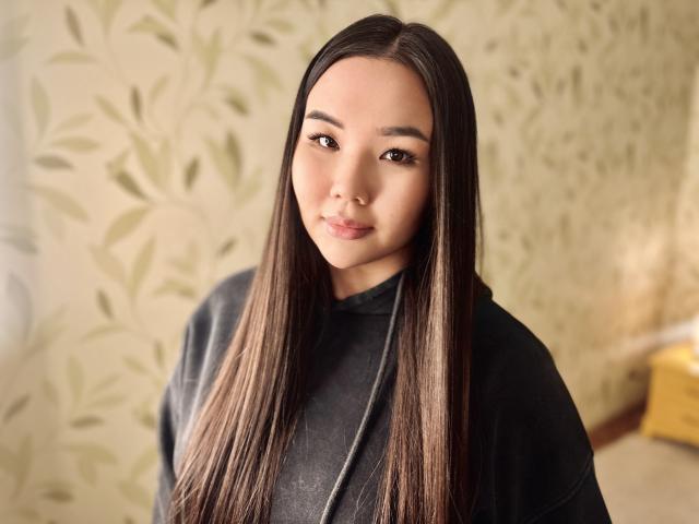 A photograph of AmandaPresle with long, straight brown hair. She has a light skin tone, brown eyes, and a subtle smile. She wears a black, textured hoodie. The background features a patterned wallpaper with green leaves. The image is well-lit with soft, natural light. AmandaPresle's hair is centered and extends past her shoulders. The texture of her hoodie contrasts with the smooth wallpaper. The composition is centered and balanced.